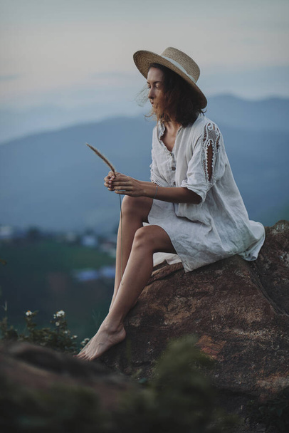 The blonde in a linen dress and straw hat sits on a viewpoint in the mountains - Foto, Imagem