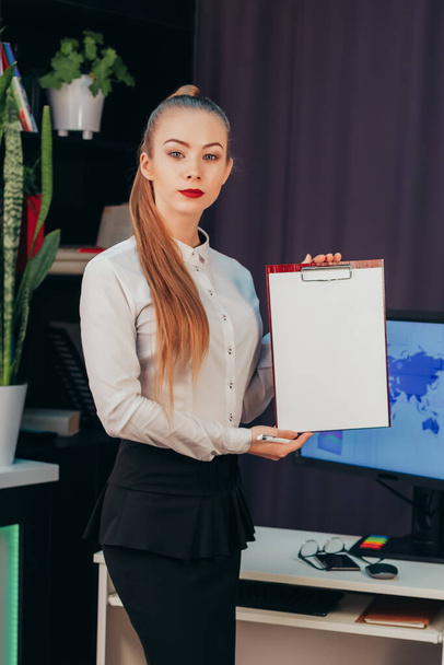 Επιχειρηματίας με μακριά μαλλιά κρατά ένα δισκίο στα χέρια της με ένα λευκό μέρος για κείμενο στο παρασκήνιο μιας επιφάνειας εργασίας με έναν υπολογιστή - Φωτογραφία, εικόνα