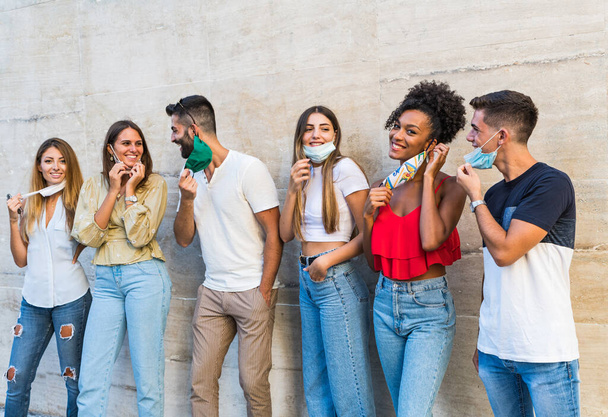 Group portrait of multi-ethnic boys and girls with colorful fashionable clothes holding friend and posing on a wall wearing face mask for coronavirus - Urban style people having fun - Concepts about youth and togetherness in covi19. - Photo, Image