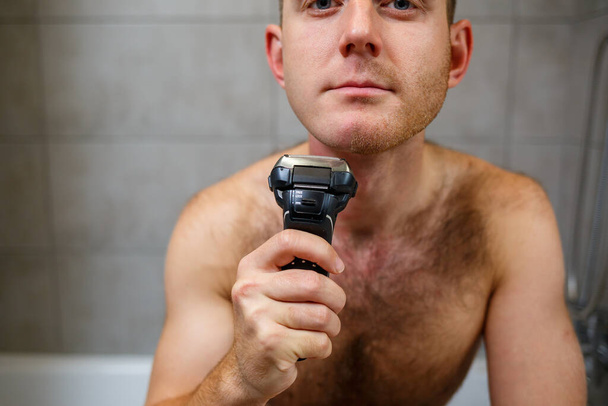 Mężczyzna goli twarz elektryczną maszynką do golenia przed lustrem. Podrażnienie skóry. Procedura kąpielowa - Zdjęcie, obraz