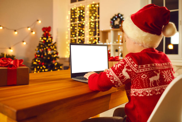 Boy in festive Santa costume and hat sitting and typing Christmas congratulation or chatting online on laptop - Photo, image
