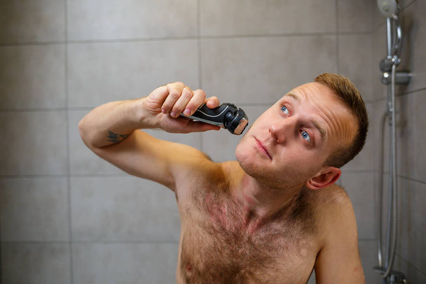 Un hombre se afeita la cara con una navaja eléctrica frente a un espejo. Irritación de la piel. Procedimiento de baño - Foto, imagen