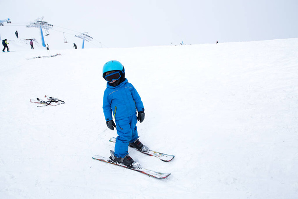 νήπιο σε κράνος, γυαλιά, σκι και μπλε φόρμες στέκεται στην χιονισμένη πλαγιά του βουνού. Μάθημα παιδικού σκι στη σχολή σκι. Χειμερινή ενεργή ψυχαγωγία για τα παιδιά, αθλητική εκπαίδευση. Ενεργή παιδική ηλικία - Φωτογραφία, εικόνα