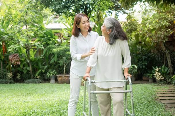 高齢のアジア系の女性は、娘と一緒にウォーカーを利用して裏庭を歩く。幸せな退職の概念介護者からのケアと貯蓄とシニア健康保険 - 写真・画像