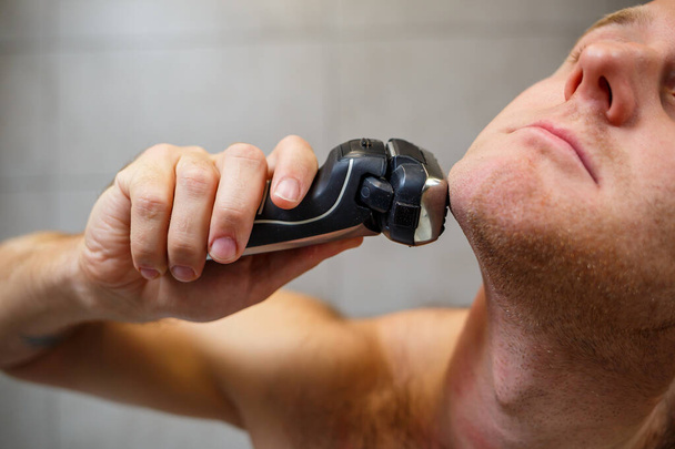 Mężczyzna goli twarz elektryczną maszynką do golenia przed lustrem. Podrażnienie skóry. Procedura kąpielowa - Zdjęcie, obraz
