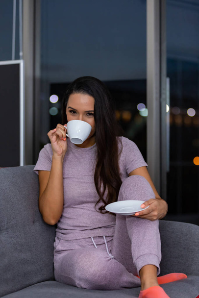 Жінка насолоджується чашкою чаю вночі, сидячи на дивані у вітальні вдома
 - Фото, зображення