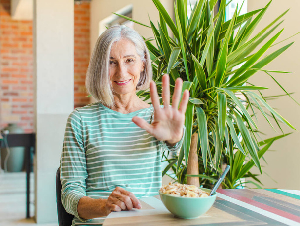 μεσήλικας γυναίκα χαμογελά και αναζητούν φιλικό, δείχνει τον αριθμό πέντε ή πέμπτη με το χέρι προς τα εμπρός, μετρώντας αντίστροφα - Φωτογραφία, εικόνα