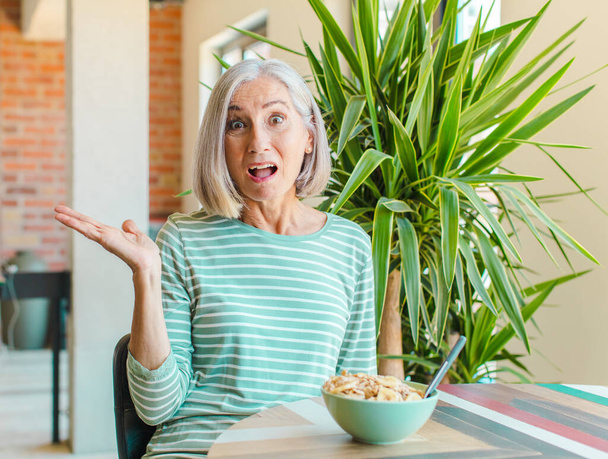 middle age woman looking surprised and shocked, with jaw dropped holding an object with an open hand on the side - Photo, Image