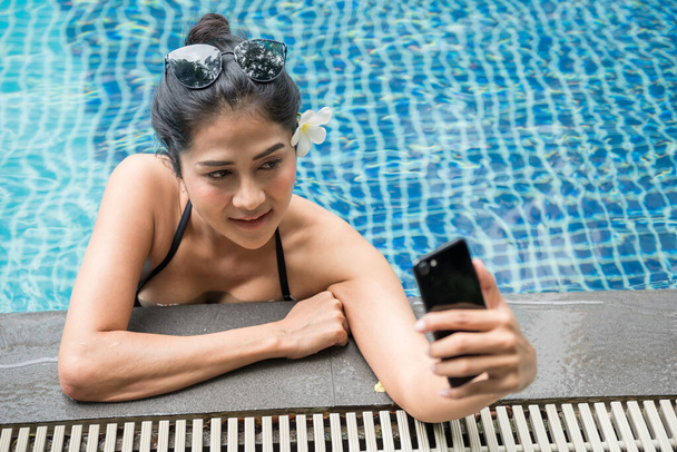 Приваблива сексуальна азіатка з чорним купальником і сонцезахисними окулярами селфі себе смартфоном на краю басейну. Прекрасна тайська дівчина відпочиває під час літніх канікул або відпусток.. - Фото, зображення