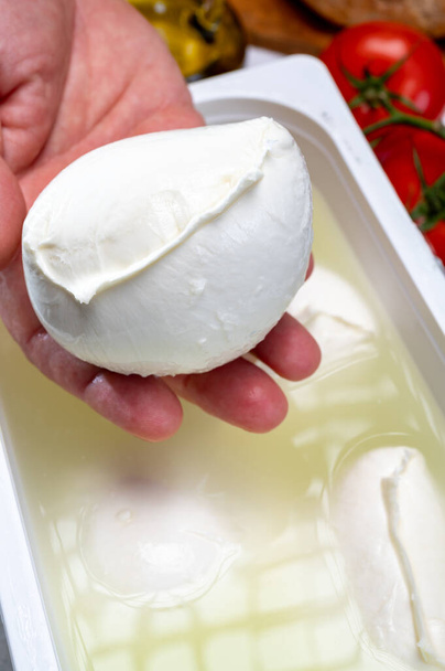 Ekspres do serów trzymający w ręku świeży ser włoski miękki z Kampanii, białe kulki sera bawolego mozzarella z mleka krowiego w pojemniku z wodą z bliska - Zdjęcie, obraz