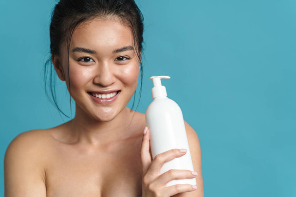 Beauty portrait of a smiling topless young asian woman posing while standing isolated over blue background, showing blank plastic bottle - 写真・画像