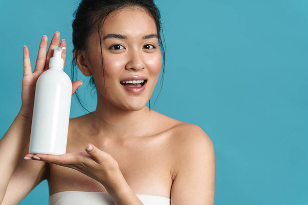 Beauty portrait of a smiling topless young asian woman posing while standing isolated over blue background, showing blank plastic bottle - Foto, Imagem