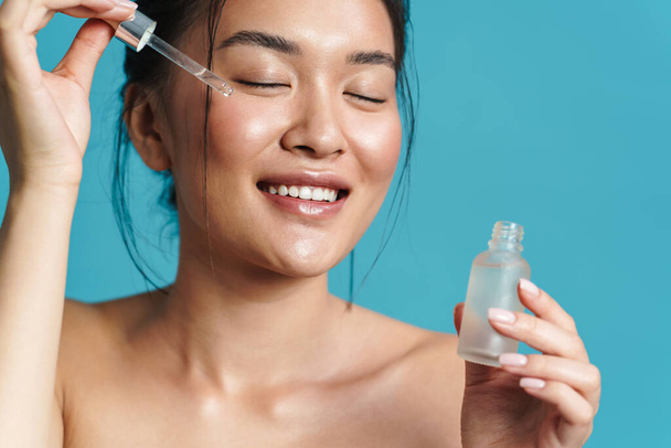 Image of happy shirtless asian girl applying facial serum isolated over blue background - Фото, изображение