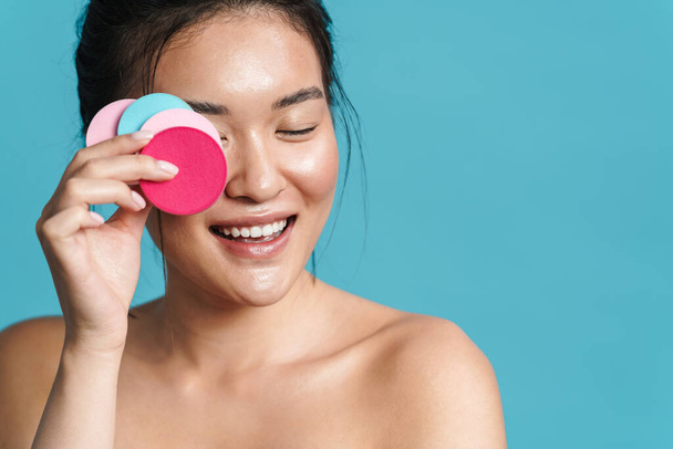 Image of joyful shirtless asian girl posing with cosmetic sponges isolated over blue background - Foto, immagini