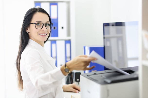 Smiling portrait of secretary who is holding papers next to the printer. - Foto, imagen