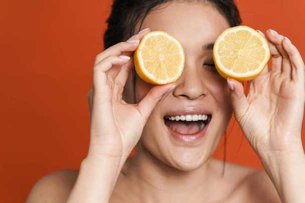 Image of shirtless asian girl laughing while posing with lemon isolated over orange background - 写真・画像