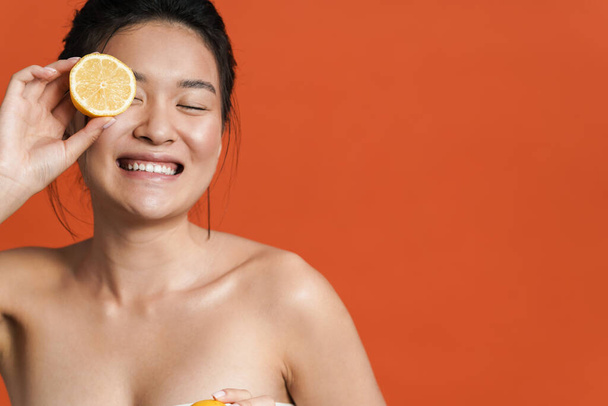Image of shirtless asian girl laughing while posing with lemon isolated over orange background - Foto, Bild
