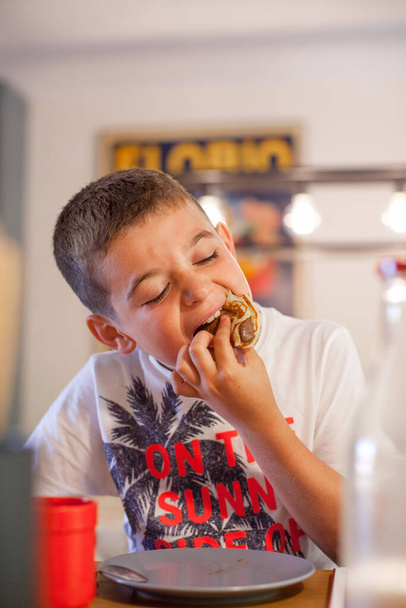 A kaukázusi fiú kinyitotta a száját, hogy a kezével egyen egy krémes csokipürével teli palacsintát. Éhes gyerek harapás reggeli egy modern konyhában reggel. - Fotó, kép