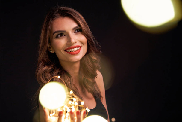 Party, holidays and celebration concept. Closeup of smiling woman holding Christmas light garland in palms, looking at camera, over dark background with bokeh - Zdjęcie, obraz