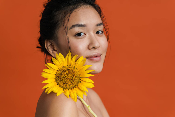Image of shirtless asian girl smiling while posing with sunflower isolated over orange background - Фото, изображение