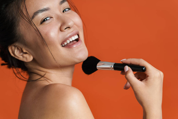 Image of cheerful shirtless asian girl making fun with powder brush isolated over orange background - Foto, afbeelding