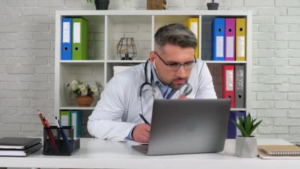 Lekarz w gabinecie szpitalnym wygląda jak szkło powiększające, pisze diagnozę w notatniku - Materiał filmowy, wideo