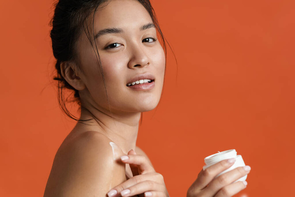 Beautiful young asian woman applying body cream isolated over orange background - 写真・画像