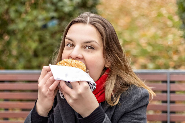Mujer joven hambrienta come hamburguesa y toma el almuerzo al aire libre en Park.Fast comida. Concepto de comida para llevar. - Foto, imagen