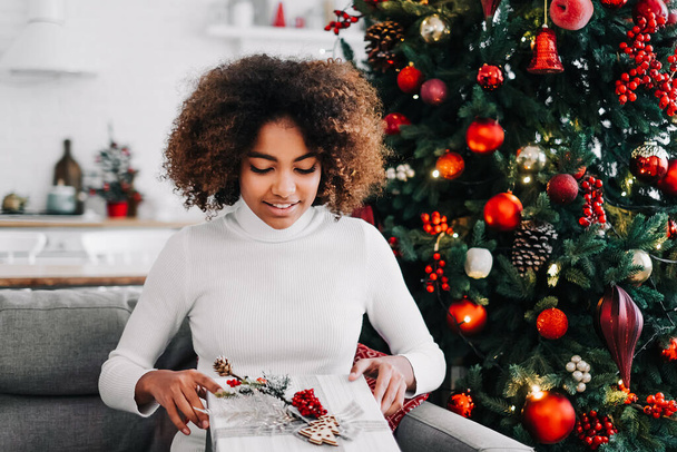 Μια όμορφη νεαρή Αφρο-Αμερικανίδα ανοίγει ένα χριστουγεννιάτικο δώρο στο σπίτι στον καναπέ στο φόντο ενός εορταστικού χριστουγεννιάτικου δέντρου. - Φωτογραφία, εικόνα
