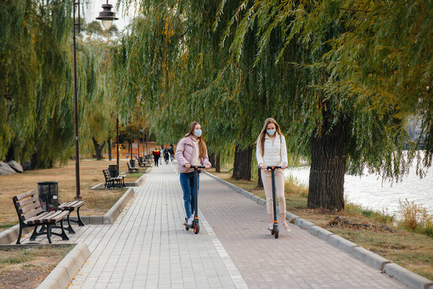 Two young beautiful girls in masks ride electric scooters in the Park on a warm autumn day. Walk in the Park. - Фото, изображение