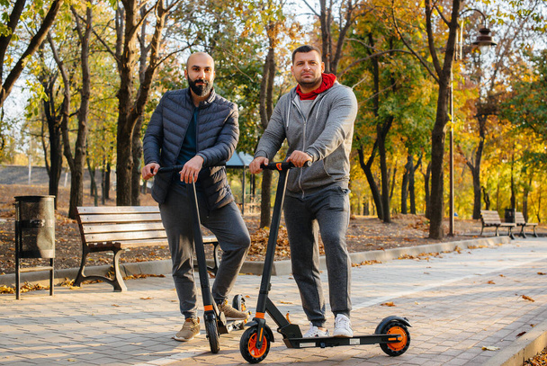 Young guys ride in the Park on an electric scooter on a warm autumn day. Walk in the Park. - Zdjęcie, obraz