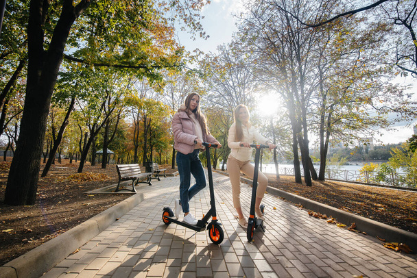 Two young beautiful girls ride electric scooters in the Park on a warm autumn day - Fotografie, Obrázek