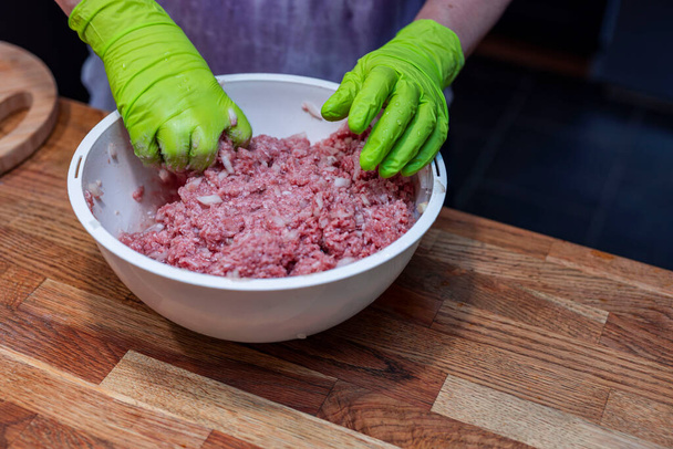 Widok żeńskich rąk w zielonych rękawiczkach pracujących z posiekanym mięsem w białej misce. Koncepcja gotowania - Zdjęcie, obraz