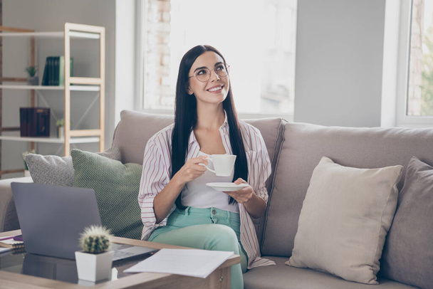 Photo of young business lady sit sofa hold mug look up dreamy imagine wear eyewear shirt in home workstation indoors - Φωτογραφία, εικόνα