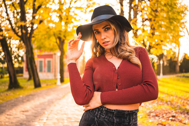 Estilo de vida otoñal al atardecer, mujer rubia caucásica en suéter rojo y sombrero negro, disfrutando de la naturaleza en un parque con árboles. Sensual mirada de chica joven mirando la cámara - Foto, imagen