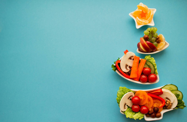 Bord in de vorm van een kerstboom met groenten, fruit, champignons en bessen. Blauwe achtergrond. Het concept van vegetarische lekkernijen voor de vakantie. Kopieerruimte. - Foto, afbeelding