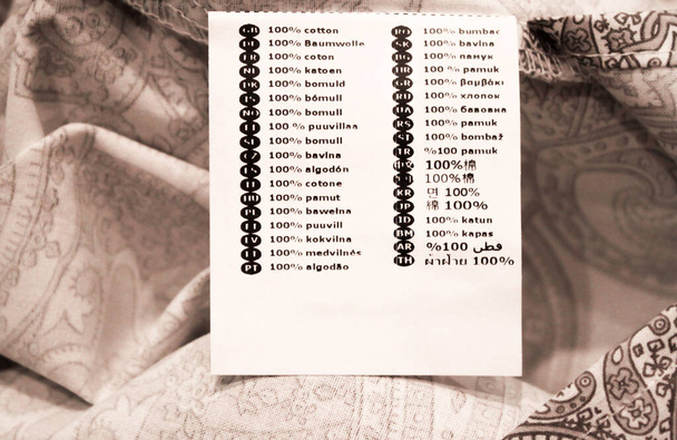 Λευκό ετικέτα πλυντήριο με πινακίδα βαμβάκι σε 36 γλώσσες στο κρεβάτι λινό. - Φωτογραφία, εικόνα