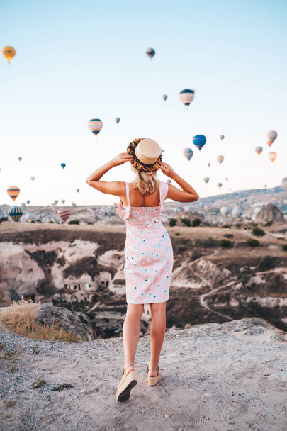 Young woman in dress and hat on a mountain top enjoying wonderful view of the sunrise and balloons in Cappadocia. Travel in Turkey concept - Φωτογραφία, εικόνα