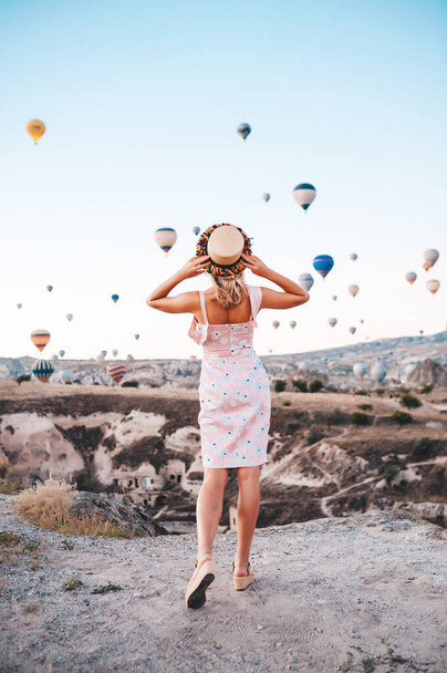 Young woman in dress and hat on a mountain top enjoying wonderful view of the sunrise and balloons in Cappadocia. Travel in Turkey concept - Фото, зображення