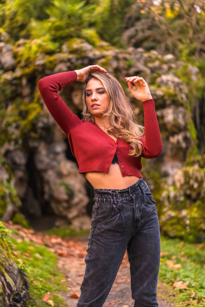 Autumn lifestyle, blonde Caucasian girl in a red sweater enjoying nature in a park. Enjoying nature with a natural cave in the background - Фото, изображение
