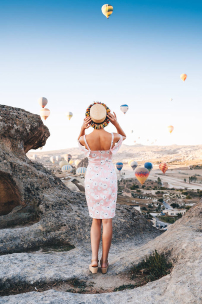 Young woman in dress and hat on a mountain top enjoying wonderful view of the sunrise and balloons in Cappadocia. Travel in Turkey concept - Foto, imagen