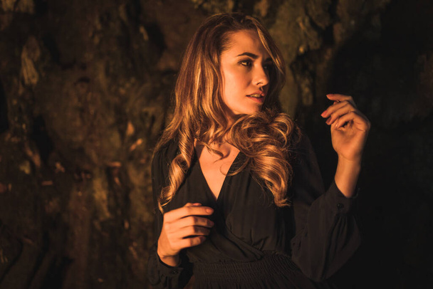 Lifestyle, a young Caucasian blonde in a black dress in a photo inside a cave, illuminated with yellow light. With the soft side light and moving the hands in the pose - Φωτογραφία, εικόνα