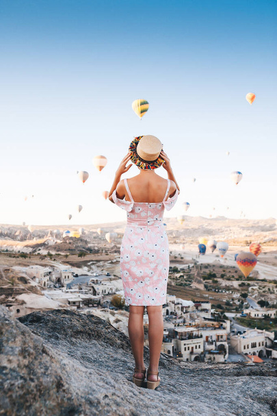 Young woman in dress and hat on a mountain top enjoying wonderful view of the sunrise and balloons in Cappadocia. Travel in Turkey concept - Valokuva, kuva