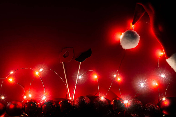 Nieuwjaarsspandoek met kerstmuts, glimmende kerstballen, snor en lippen op stokken en slinger van lichtjes op een rode achtergrond. Briefkaart met een magische vakantiesfeer en een vrolijke stemming. - Foto, afbeelding