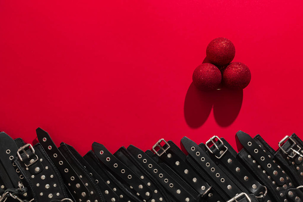 Rotes Banner mit drei Weihnachtskugeln und schwarzen Lederarmbändern und Metallnieten zum Fesseln von Händen oder Beinen und sexuellen Spielen. BDSM-Thema. Hintergrund für Werbung für Erwachsenenspielzeug oder Sexshop. - Foto, Bild