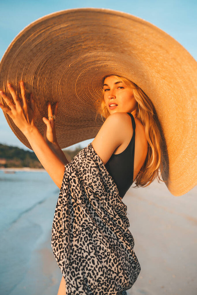 Portrait of a woman posing on the beach. Attractive female wearing black swimsuit and big fashionable hat. - Foto, afbeelding