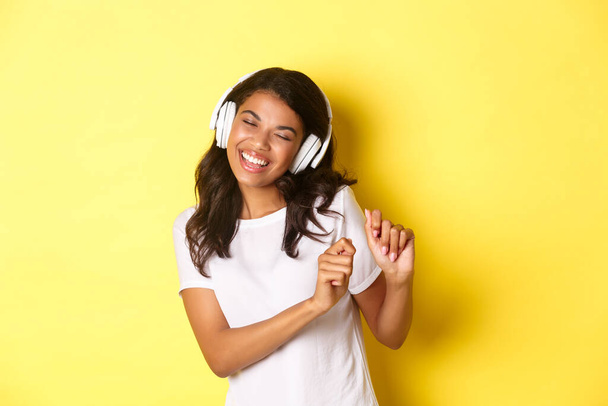 Cheerful teenage african-american girl listening to music in headphones, dancing upbeat and smiling, standing over yellow background - Photo, Image