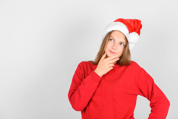 Adorable smiling girl with Santa red hat wearing red pullover and thinking of Christmas wish, dreams of gift. Christmas background - Fotoğraf, Görsel