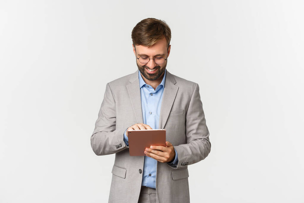 Immagine di uomo d'affari che lavora, guarda tablet digitale e sorride soddisfatto, in piedi in abito grigio e occhiali su sfondo bianco - Foto, immagini