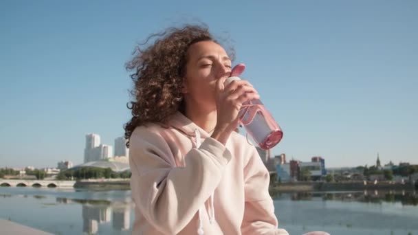 Primer plano de la joven mujer caucásica bebiendo agua de la botella de plástico deportivo que tiene descanso después de un duro entrenamiento al aire libre con pintoresco horizonte con cielo azul y río en el fondo - Imágenes, Vídeo